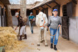Elderly woman horse farm owner leading white racehorse along stalls outdoor while Asian female worker stacking hay with pitchfork in yard. Daily chores in stable