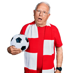 Poster - Senior man with grey hair football hooligan holding ball in shock face, looking skeptical and sarcastic, surprised with open mouth