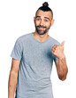 Hispanic man with ponytail wearing casual grey t shirt smiling with happy face looking and pointing to the side with thumb up.