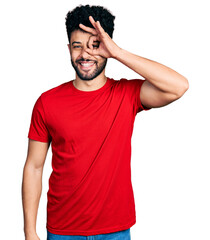 Canvas Print - Young arab man with beard wearing casual red t shirt doing ok gesture with hand smiling, eye looking through fingers with happy face.