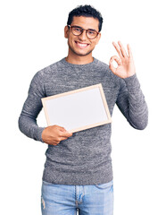 Wall Mural - Hispanic handsome young man holding blank empty banner doing ok sign with fingers, smiling friendly gesturing excellent symbol