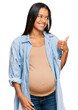 © Krakenimages.com - Beautiful hispanic woman expecting a baby showing pregnant belly smiling with happy face looking and pointing to the side with thumb up.