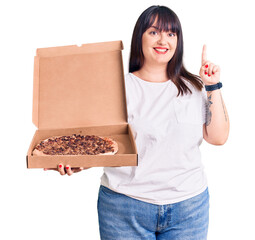 Sticker - Young plus size woman holding delivery pizza box surprised with an idea or question pointing finger with happy face, number one