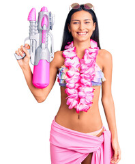 Poster - Young beautiful latin girl wearing bikini and hawaiian lei holding water gun looking positive and happy standing and smiling with a confident smile showing teeth