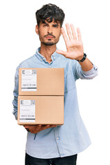 Wall Mural - Young hispanic man holding delivery package with open hand doing stop sign with serious and confident expression, defense gesture