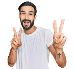 Wall Mural - Young hispanic man wearing casual white t shirt smiling looking to the camera showing fingers doing victory sign. number two.