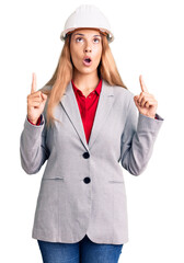 Poster - Beautiful young woman wearing architect hardhat amazed and surprised looking up and pointing with fingers and raised arms.