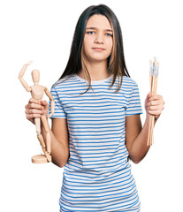 Sticker - Young brunette girl with long hair holding small wooden manikin and pencils clueless and confused expression. doubt concept.