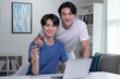 © amenic181 - Young Asian gay couple smiling looking at camera while holding credit card and using laptop computer. LGBT gay businessman working at home. Online shopping, e-commerce, internet banking.