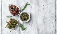 Caper Berries And Olives On White Plate With Fresh Basil Leaves On A Gray Background. Mediterranean Snack Assortment. Banner, Menu, Recipe Place For Text, Top View