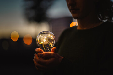 young worried female human holding bright shining lightbulb in her hands feeling unsave while thinking negative about a future blackout with no electricity or power outage with warm background