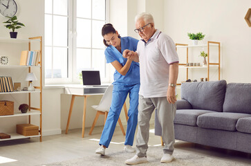 Young nurse helping elderly man walk in the room, holding his hand, supporting him. Treatment and rehabilitation after injury or stroke, life in assisted living facility, senior care concept