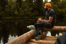 An African American Man With An Orange Beanie Sitting On A Wooden Raft On A Wide River Holding A Fishing Rod.