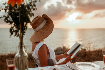 Wall Mural - a young woman in a hat sits by the sea at sunset, reads a book and drinks champagne