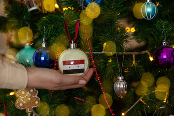 Close up of girls hand holding a Christmas ball for a fir tree with the flag of Syria. New year in Syria. New year holiday greeting card with copy space for text. Decorating home for celebration.