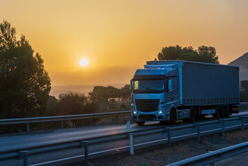 Truck with a tarpaulin semi-trailer driving on a highway with the sun in the background at dawn.