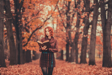 Wall Mural - Beautiful girl with red hair in a sweater reads a book in the autumn park, October