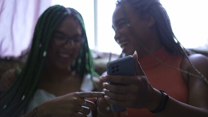 Wall Mural - Girlfriends pointing at cellphone screen laughing and smiling. Two young female friends holding phone