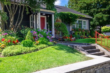 The front yard of a dark blue house with a vibrant red door. The building has a glass window with white trim. The colorful garden has green grass, red, pink, and yellow flowers, shrubs, and trees. 