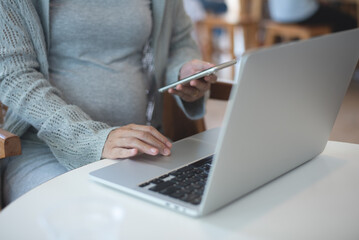 Canvas Print - Pregnant business woman sitting at table using mobile phone and working on laptop computer at cafe. pregnant woman online shopping via mobile app, close up