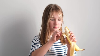 A little hungry girl eats a ripe yellow banana on a white background. The girl bites a banana