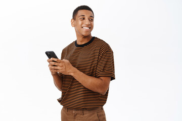 Wall Mural - Mobile technology concept. Smiling african american man using his smartphone, holding phone and looking happy, texting in application, white background