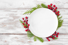 Christmas Or Winter Table Setting Mock Up. Blank White Plate With Frame Of Evergreen Branches And Red Berries. Above View On A White Wood Background. Copy Space.