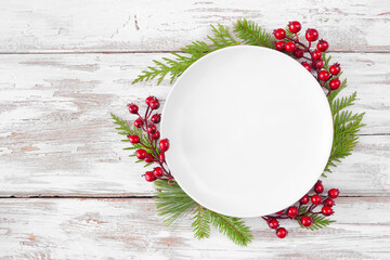 Wall Mural - Christmas or winter table setting mock up. Blank white plate with frame of evergreen branches and red berries. Above view on a white wood background. Copy space.
