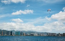 Person Paragliding Over Water With A Cityscape And Clouds In The Background