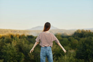 A young woman stands with her back to the camera with her hands up in a T-shirt and jeans in nature and enjoys a beautiful view of the mountains. Autumn travel to nature lifestyle