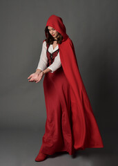 Full length portrait of beautiful brunette woman wearing red medieval fantasy costume with long skirt and flowing hooded cloak.
Standing pose with gestural hand poses, isolated on grey studio backgrou