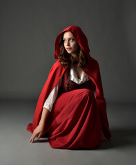 Wall Mural - Full length portrait of beautiful brunette woman wearing red medieval fantasy costume with long skirt &  flowing hooded cloak.
Sitting pose with gestural hand pose, isolated on grey studio background