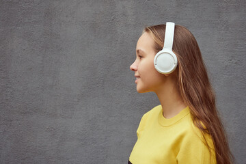 profile view of teenage girl in yellow sweater listening to music in wireless headphones on gray background