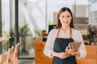 © Kritdanai - Opening a small business, AHappy Asian woman in an apron standing near a bar counter coffee shop, Small business owner, restaurant, barista, cafe, Online, SME, entrepreneur, and seller concept