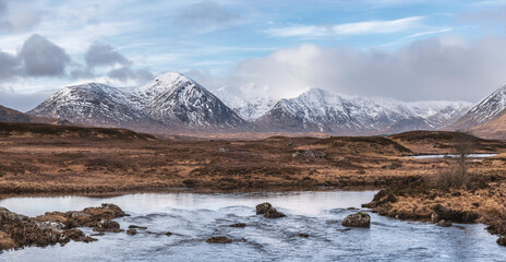 Wall Mural - Majestic Winter panorama landscape image of mountain range and peaks viewed from Loch Ba in Scottish Highlands with dramatic clouds overhead