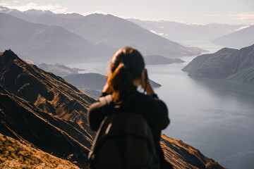 caucasian girl from the back with black t-shirt and backpack on her back calm relaxed photographing the beautiful landscape of big mountains and big calm lake, roys peak, new zealand