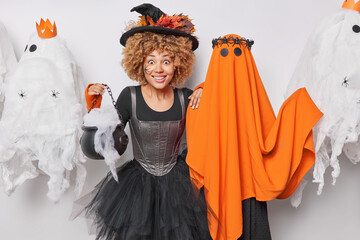 Wall Mural - Surprised cheerful woman witch poses cauldron smiles gladfully wears black hat and dress stands near spooky ghost enjoys party on Halloween night poses around spooky creatures. 31st of October