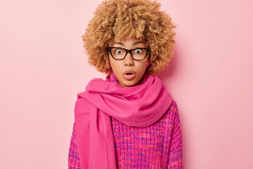 Wall Mural - Portrait of surprised shocked woman with blonde curly hair stares through transparent eyeglasses wears knitted jumper and scarf around neck isolated on pink background. Omg and human reactions concept