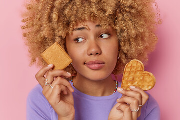 Wall Mural - Headshot of curly haired sad young woman chooses between cookie and heart shaped waffle has sweet tooth looks aside dressed in casual clothes isolated over pink background. Breaking diet concept