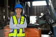 African American female driver forklift truck in heavy metal industrial factory. Smiling woman engineer wearing vest and helmet safety standing and arms crossed at warehouse factory.