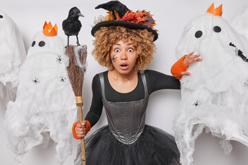 Wall Mural - Photo of shocked surprised woman with curly hair stands speechless holds broom crow on it celebrates Halloween poses around creepy creatures prepares for mysterious event preapares for spooky event