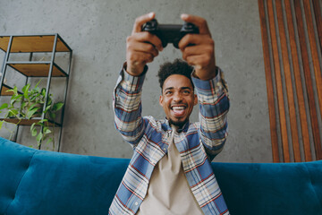 Bottom view young man in shirt sit on blue sofa couch hold in hand play pc game with joystick console do winner gesture stay at home hotel flat rest relax spend free spare time in living room indoor