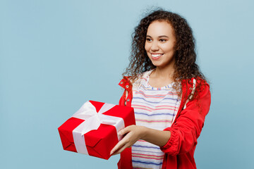 Wall Mural - Side view young woman of African American ethnicity wears red jacket hold giving present box with gift ribbon bow isolated on plain pastel light blue cyan background Wet fall weather season concept.