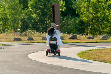 Woman with a disability in the wheelchair mobility scooter enjoying summer park in the evening. Carefree woman with mobility issue uses electric three wheel wheelchair scooter having fun.