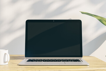 Poster - designer office desktop with empty laptop display with mock up place in frame, concrete wall background with sunlight and shadows, blurry decorative plant and coffee cup. 3D Rendering.