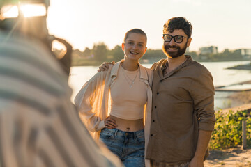 Wall Mural - Photographer POV. Young caucasian couple asking someone to take their picture. Bald queer person in their 20s posing with caucasian bearded hipster guy by the river. Blurred unrecognizable