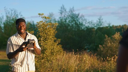 Wall Mural - Photography as a way to save memories. Tall handsome black guy holding old instant analog camera while standing in nature. Blurred person silhouette in the foreground. . High quality 4k footage