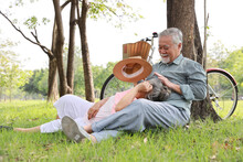 Portrait Of Happy Asian Senior Man And Woman Lying On Lap And Embracing With Bicycle And Hat In Summer Garden Outdoor. Lover Couple Going To Picnic At The Park. Happiness Marriage Lifestyle Concept.