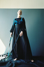 Portrait Of Fashion Model With Shaved Head In Long Transparent Dress Standing Against Blue Wall
