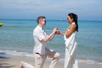 Surprised man kneeling holding a ring to propose to his girlfriend on the beach bright blue sea.